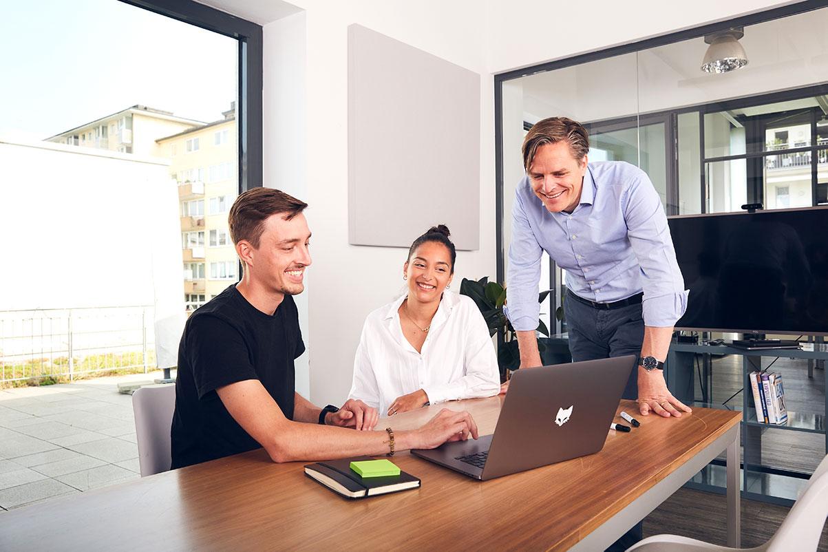 Three coworkers (two sitting, one standing) look smilingly into a laptop as they work in a bright office conference room.